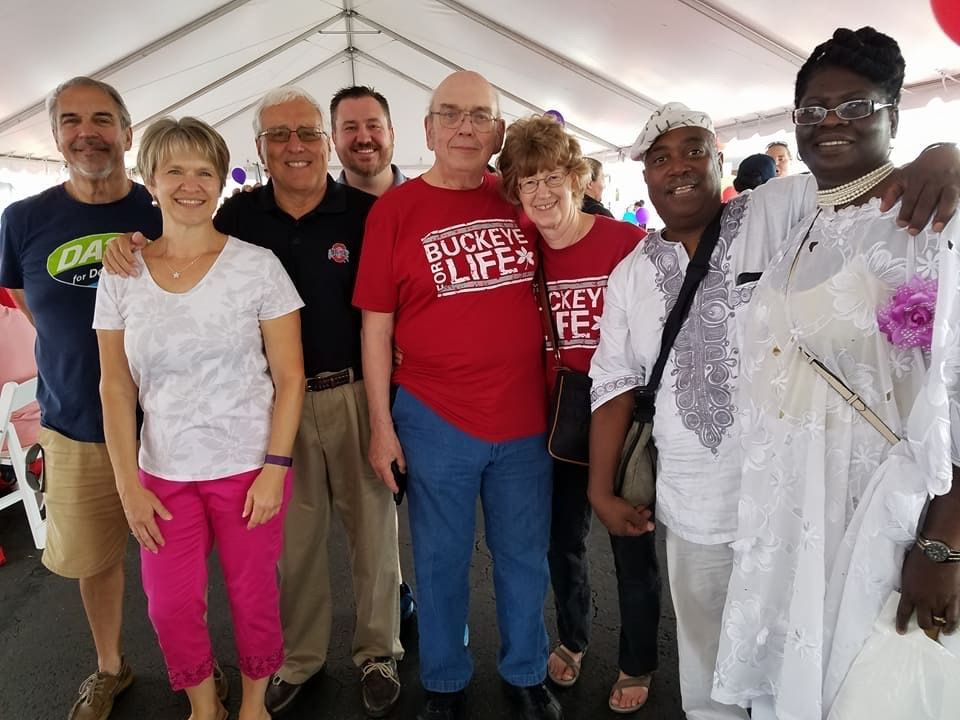 Group of people smiling, posing for photo under a white tent. They are of diverse ages and ethnicities.