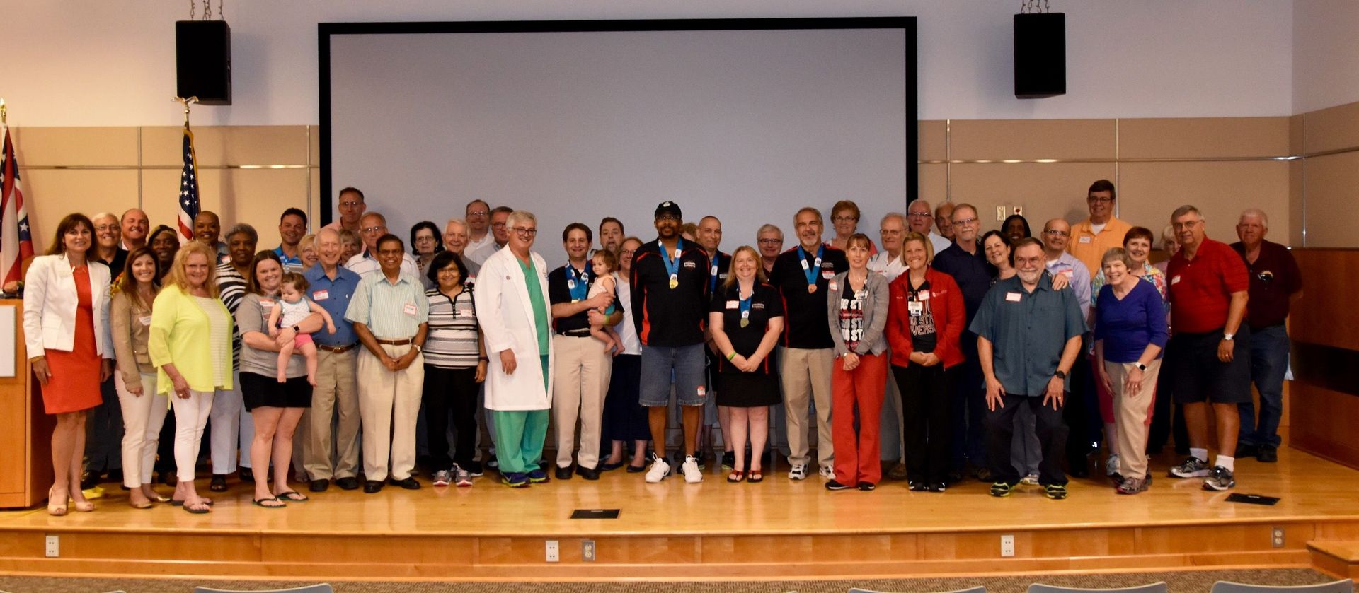 Group of people on a stage in front of a screen. People are smiling, some are wearing medals, indoors.