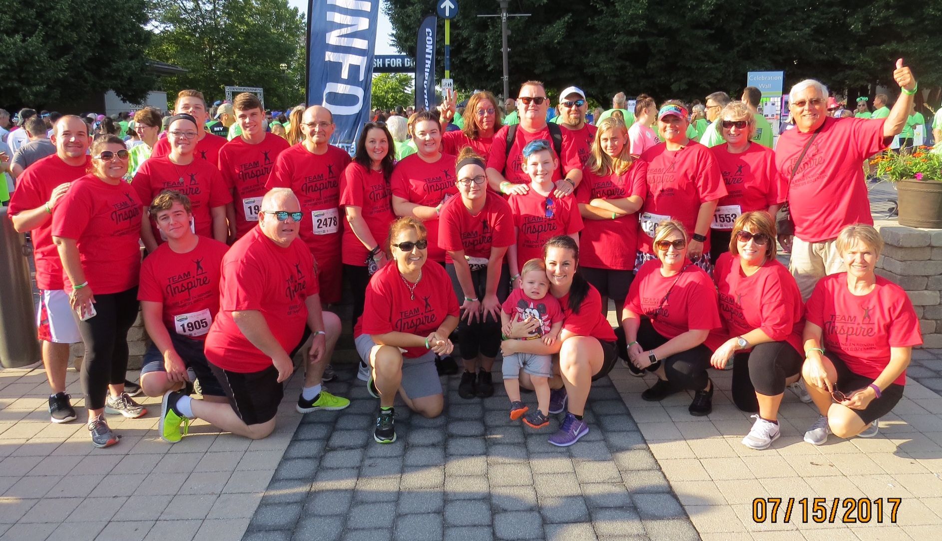 Group of people in red shirts at an outdoor event, smiling. Some are holding babies. July 15, 2017.