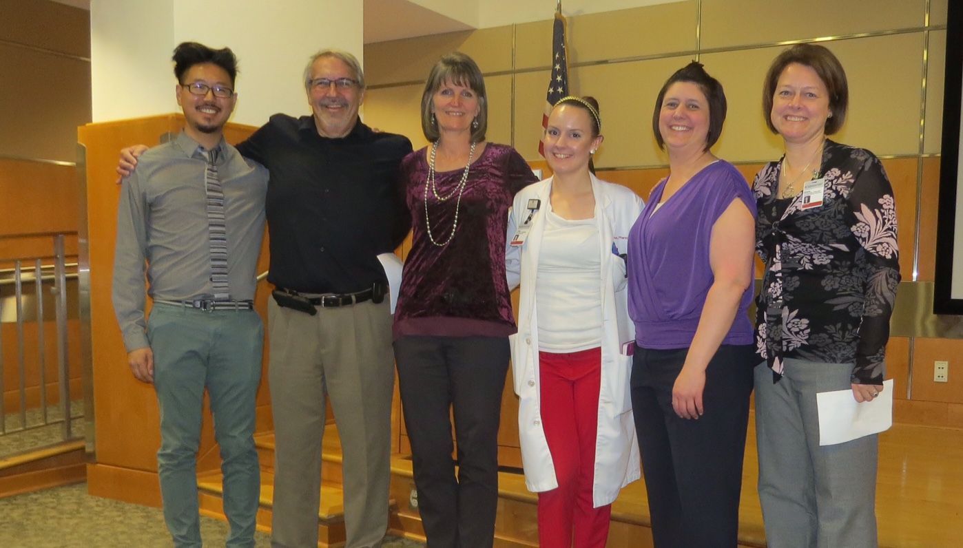 Group of six people, arms around each other, posing. Indoors, various outfits, smiles, wood paneling in background.