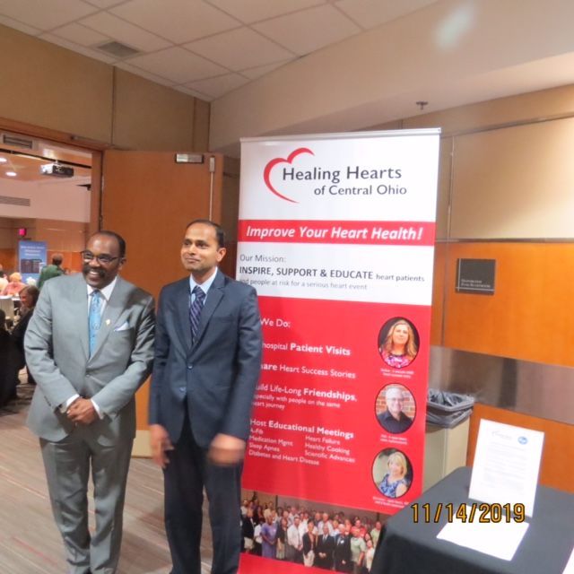 Two men in suits stand by a "Healing Hearts" banner; an event setting.