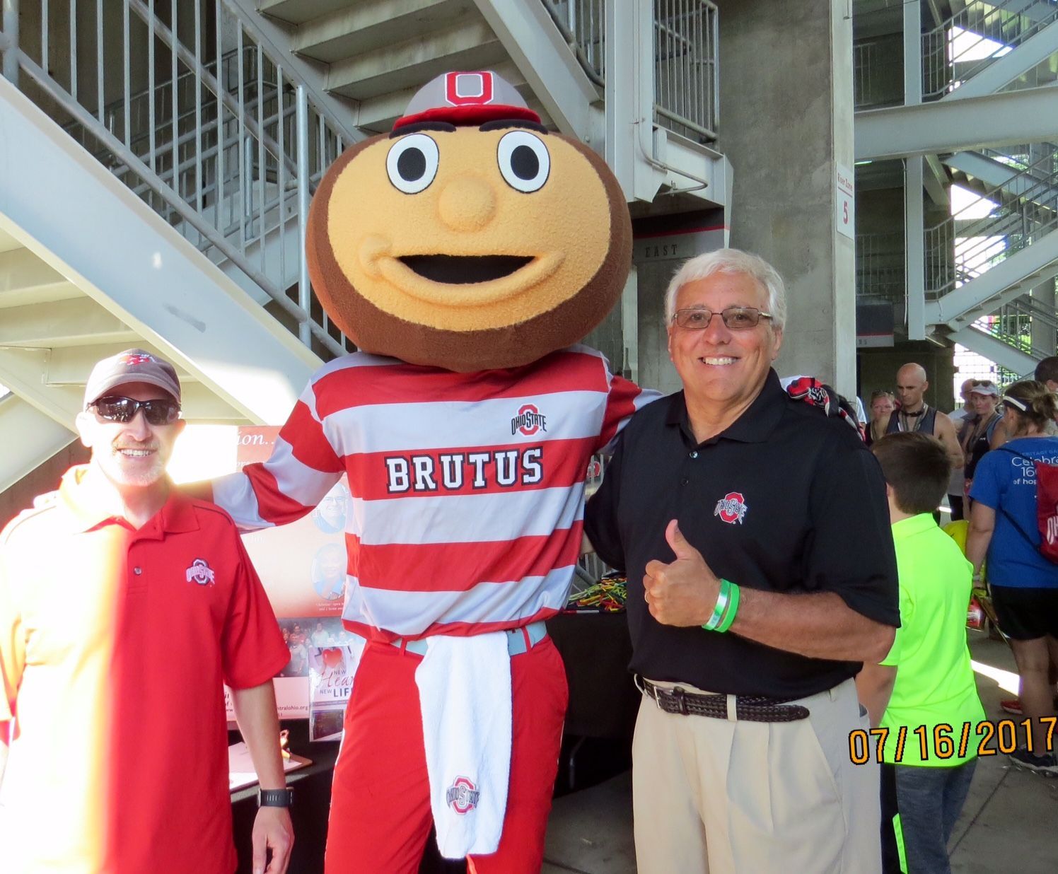 Brutus Buckeye with two men. Brutus in red/white with arms around the men. Outdoors, 2017.