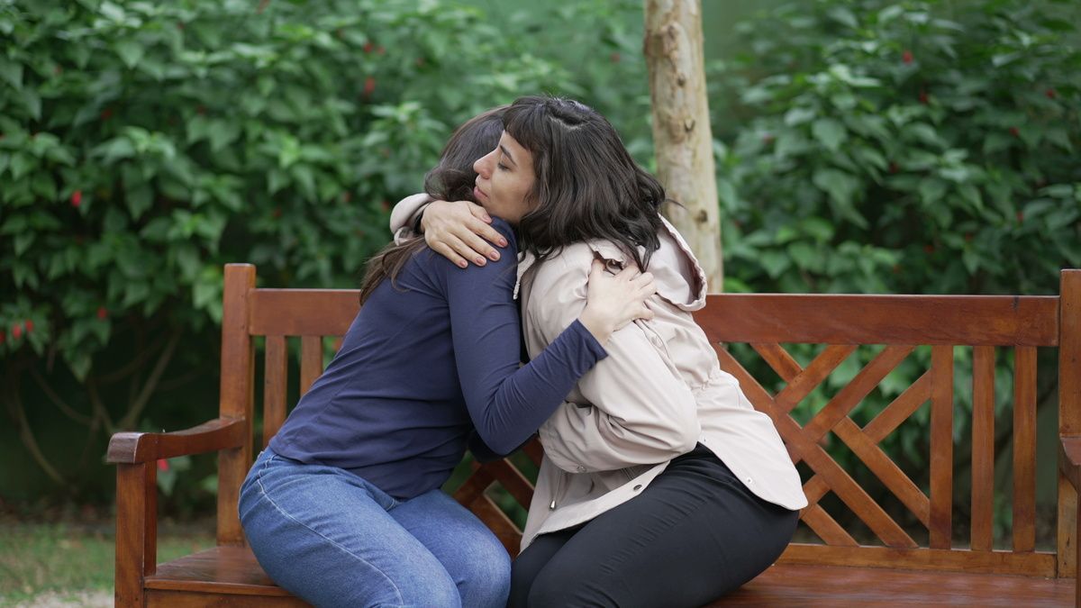 Two people embracing on a park bench. One in a blue shirt, the other in a tan jacket. Green foliage in the background.