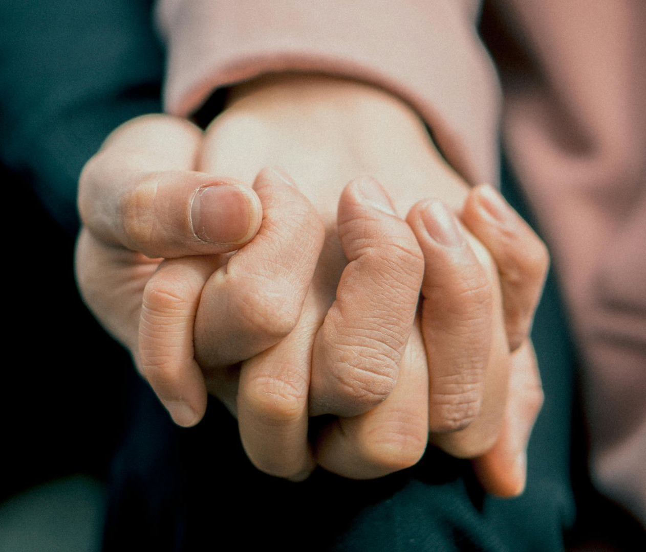 Hands clasped, one hand on top. A light pink sleeve is visible in the background.