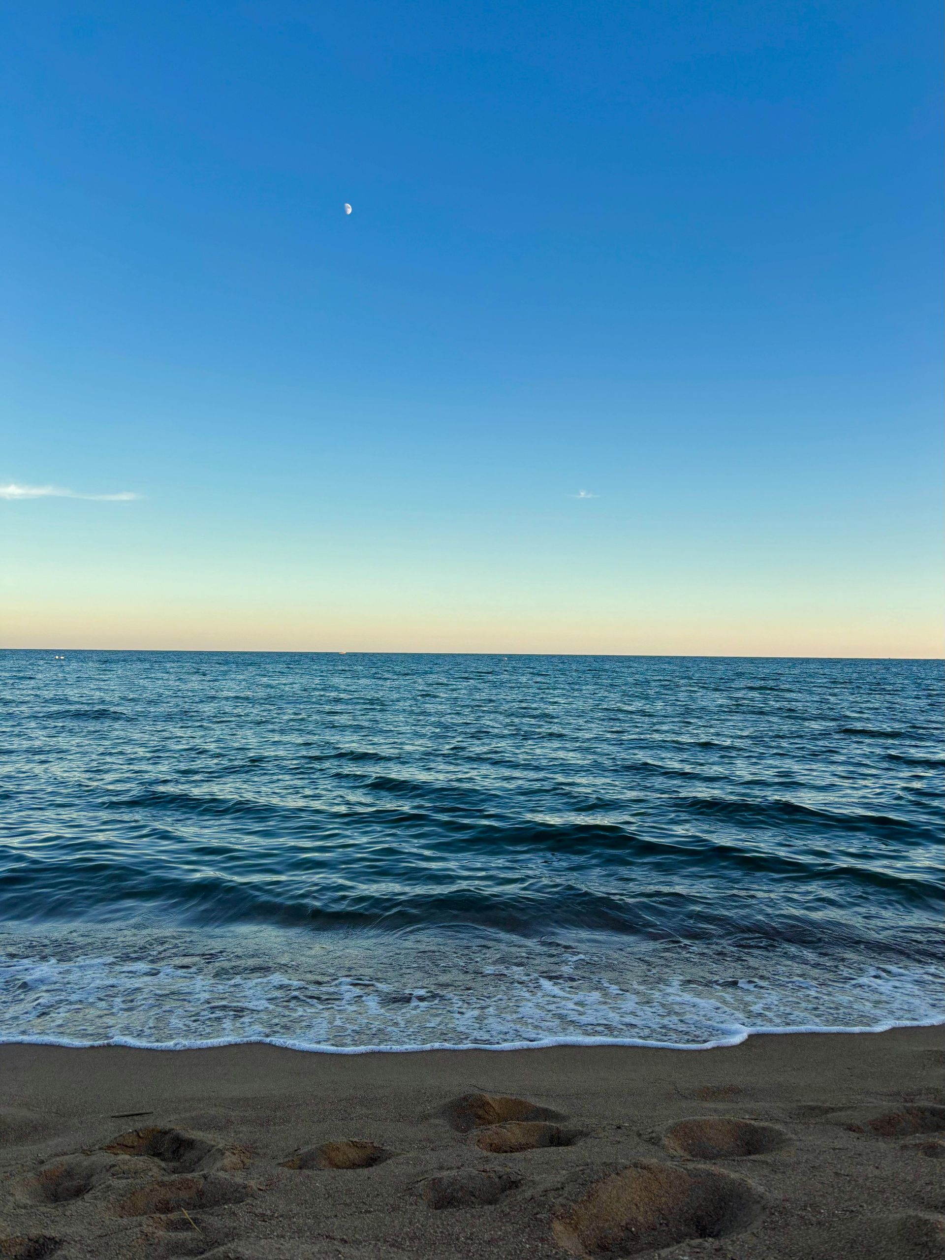 Ocean waves rolling onto a sandy beach under a clear, blue sky with a crescent moon.