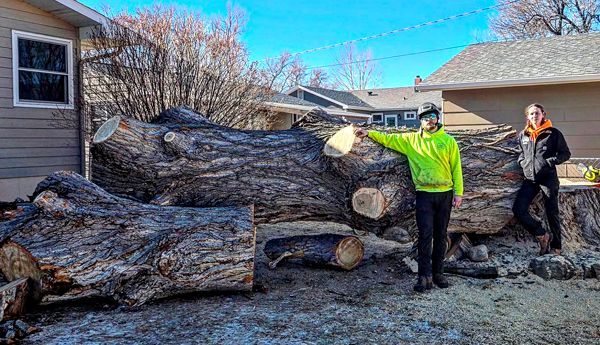Two people are standing next to a large log in front of a house.