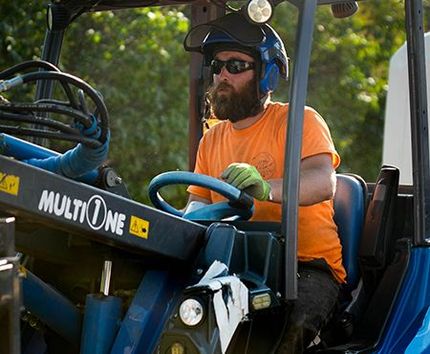 Man driving a blue MultiOne tractor, wearing safety glasses and a hard hat. Outdoors in daylight.