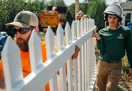 Two men installing a white picket fence; one wears a helmet and green shirt, the other an orange shirt and sunglasses.