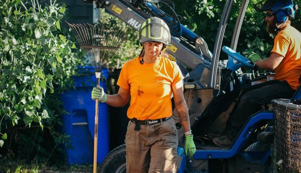Woman in orange shirt and helmet holding rake near small blue tractor; outdoors.