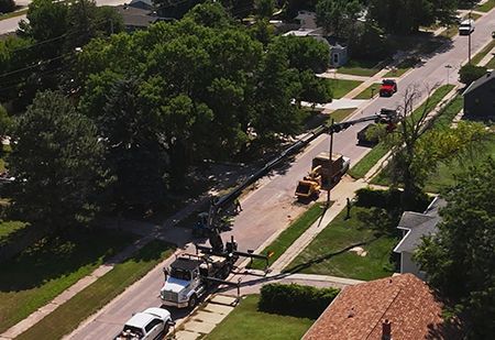 A crane lifts a tree section over a road in a residential area; a truck and other vehicles are present.