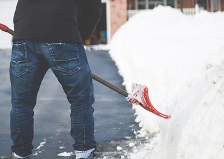 Person shoveling snow from a driveway; red shovel, dark blue jeans, black jacket.