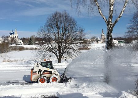 Bobcat snowblower clearing snow on a bright, sunny day; buildings and trees in background.