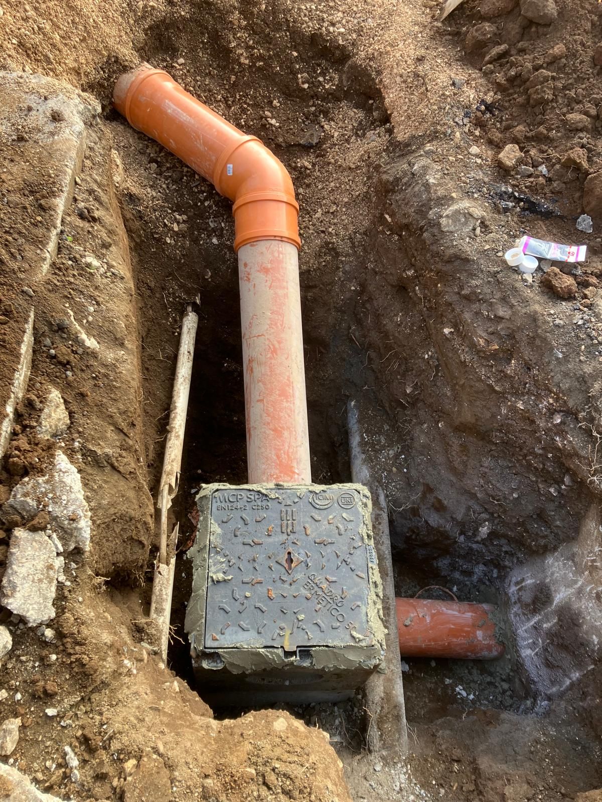 Tubo in PVC arancione interrato e scatola di derivazione quadrata in una trincea scavata nel terreno.