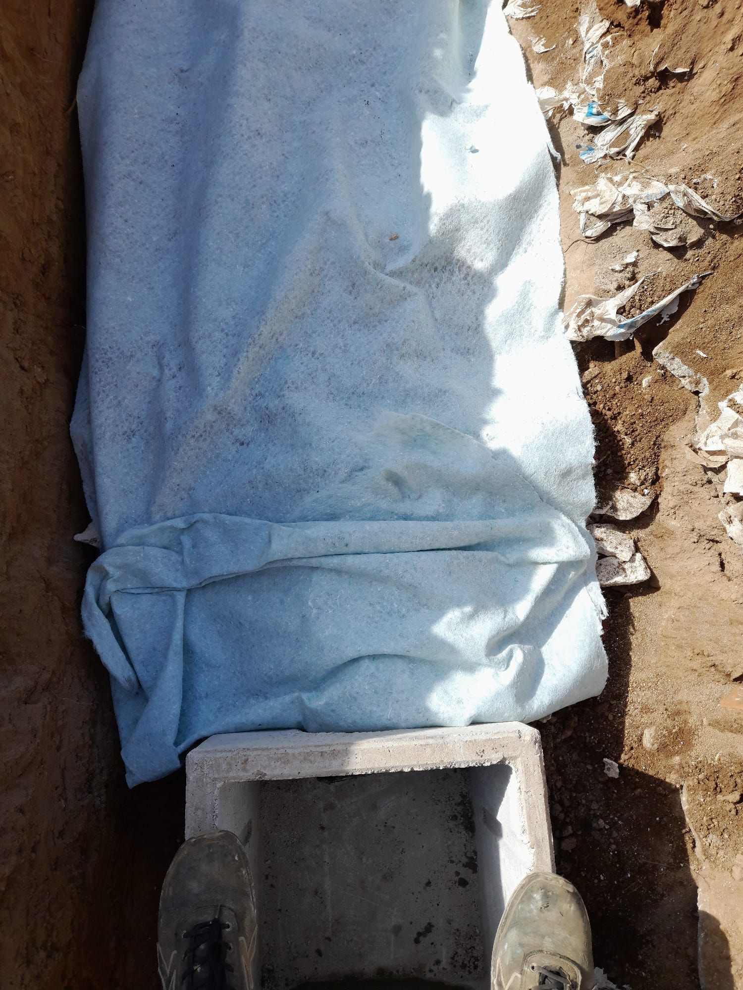Tubo di scarico coperto da un telo blu accanto a un blocco di cemento in una trincea di terra.