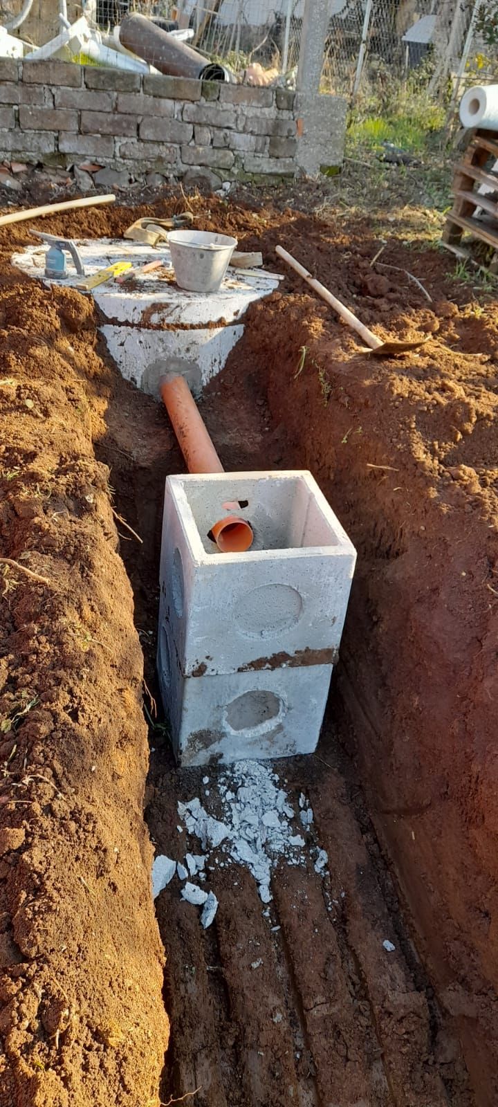 Tubazione di servizio interrata con blocchi di cemento e tubo protettivo rosso in un cantiere edile all'aperto.