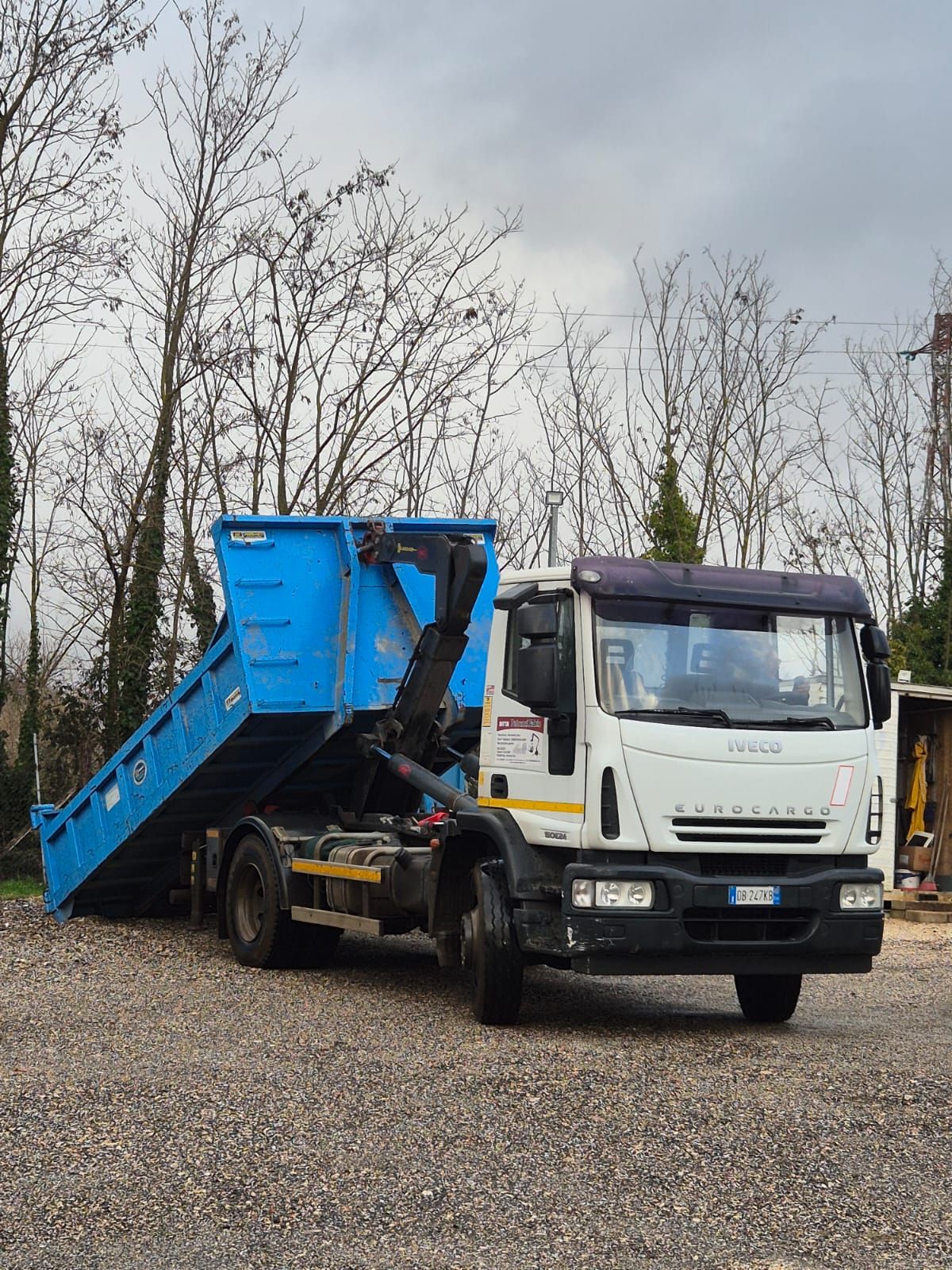 Camion della spazzatura blu e bianco parcheggiato su ghiaia con alberi spogli sullo sfondo.