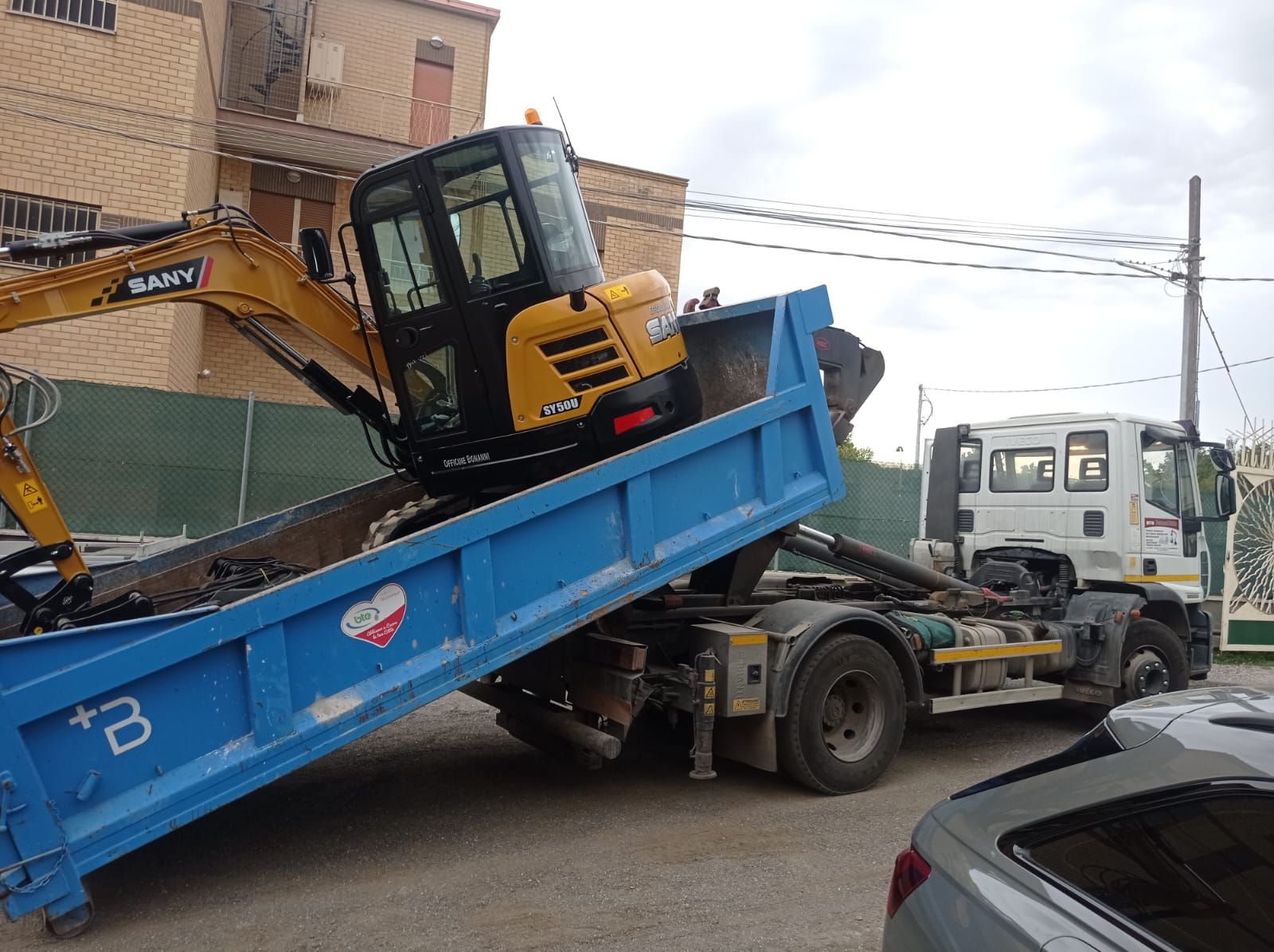 Un carro attrezzi solleva un rimorchio ribaltabile blu con un escavatore giallo sul luogo di un incidente stradale.
