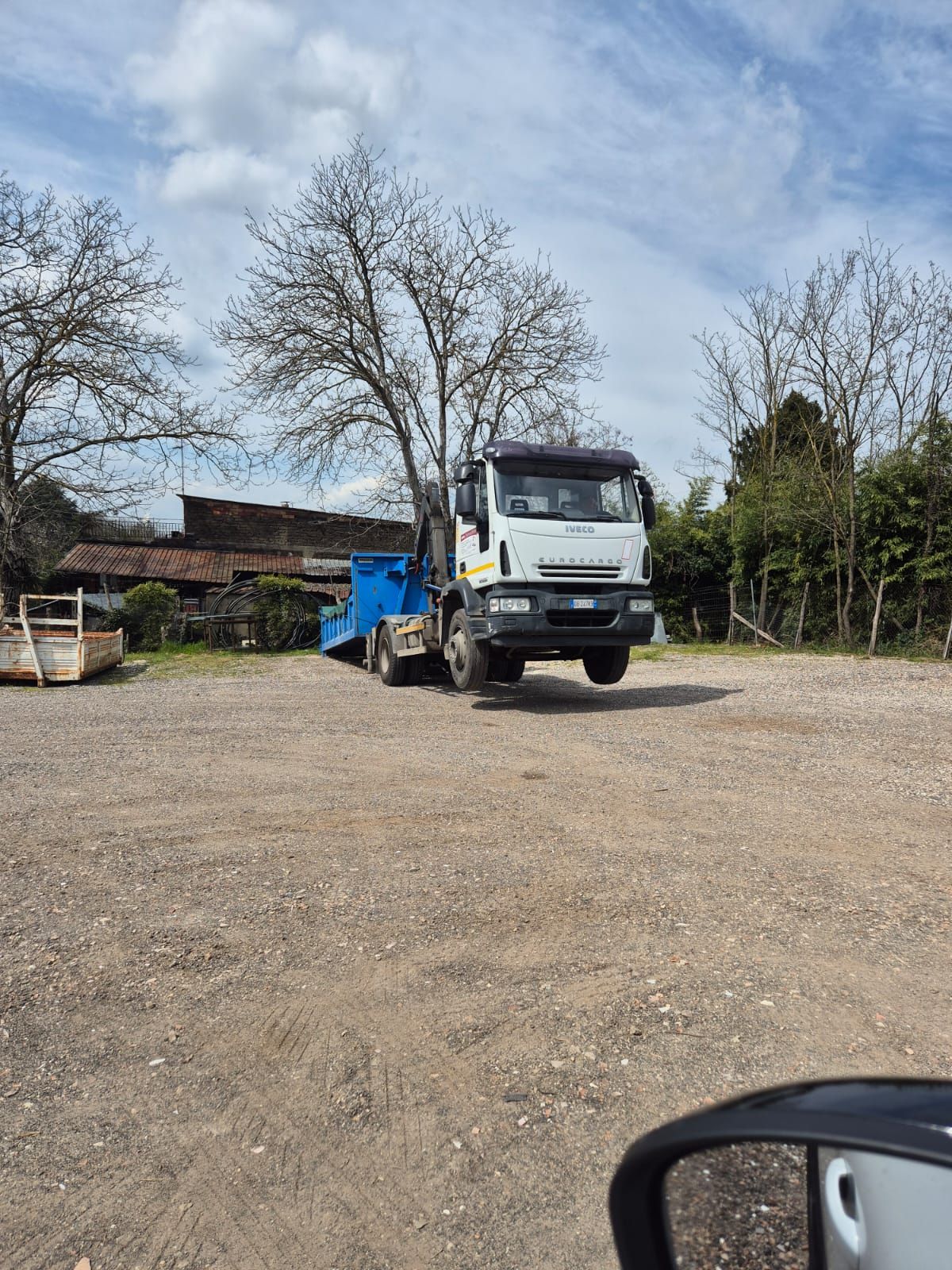 Autocarro ribaltabile bianco parcheggiato su un piazzale ghiaioso con alberi ed edifici sullo sfondo.