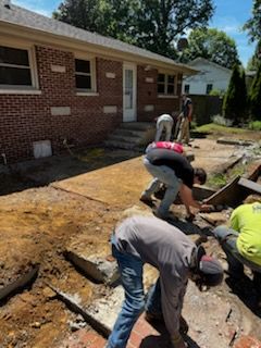 Workers digging up a cracked driveway beside a brick house on a sunny day