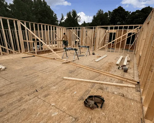 A construction worker frames the wooden wall structure of a house on a plywood subfloor under a sunny sky.