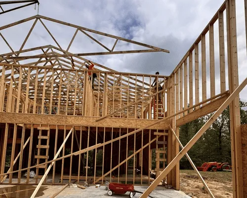 Construction workers frame the wooden structure and roof trusses of a two-story building under a cloudy sky.