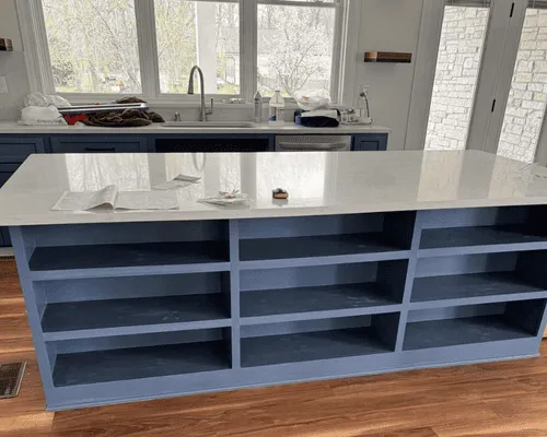 A kitchen island with a white countertop and blue open shelving, situated in a room with hardwood floors and windows.