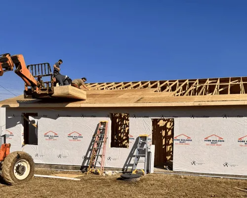 Construction workers on a forklift platform install roof sheathing on a house frame against a clear blue sky.
