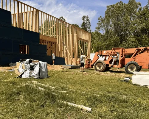 Construction site with a wooden building frame, a partially sheathed wall, two people, and an orange telehandler.