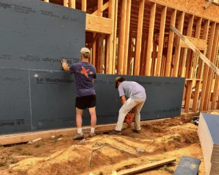 Two workers install grey construction panels onto the wooden frame of a building under construction.