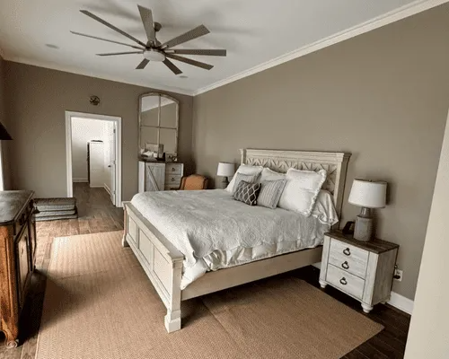 A bedroom features a white wooden bed with grey bedding, two bedside tables with lamps, and a ceiling fan on beige walls.
