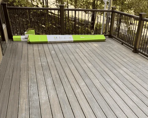 A gray composite deck with a dark metal railing, featuring a long green and white box of building materials on the floor.