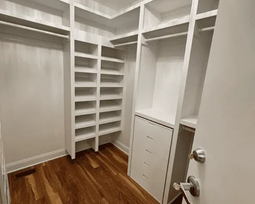 A walk-in closet featuring white wooden shelving, vertical cubbies for shoes, and a set of four drawers on hardwood floors.