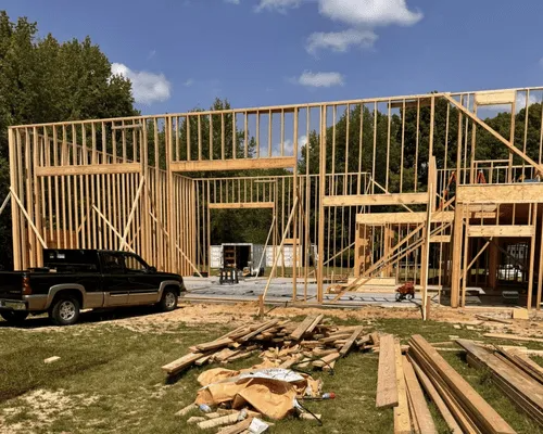 A wood-framed house under construction on a sunny day with a black pickup truck parked in the foreground.