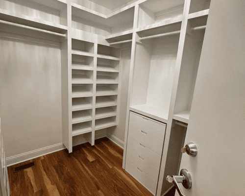 A walk-in closet featuring white wooden shelving, clothing rods, and a four-drawer unit over polished hardwood flooring.