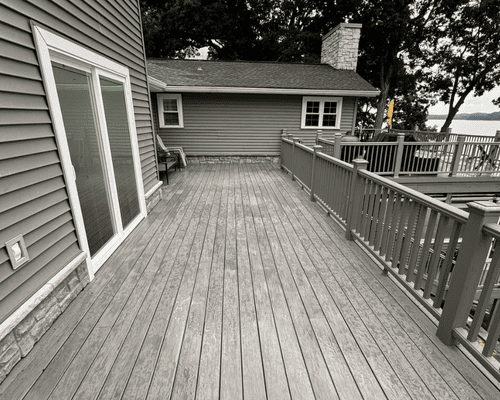 A wooden deck with gray railings extends from a house, overlooking a lake with trees in the background.