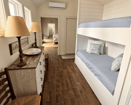 A bedroom with white bunk beds, wooden floors, a dresser, and two table lamps, leading into an adjacent room.
