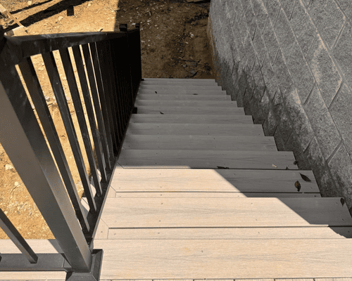Top-down view of light-gray composite stairs with a black metal handrail, leading down to dirt beside a cinder block wall.