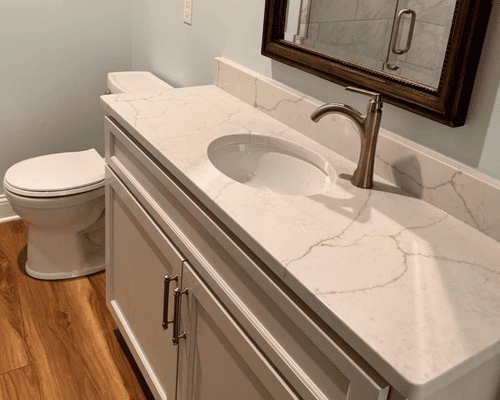 A bathroom vanity with a white marble-look countertop, a bronze faucet, and a white toilet on a wood-look floor.