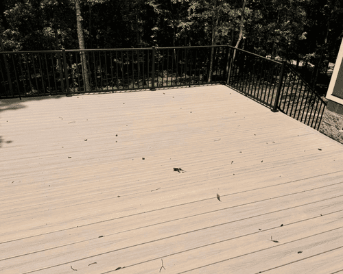 A light tan composite deck with a black metal railing, surrounded by trees on a sunny day.