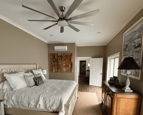 A cozy bedroom featuring a large white bed, a ceiling fan, a wall-mounted AC unit, and a dresser with a lamp.