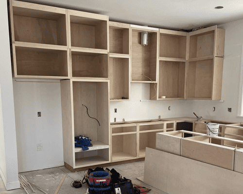 Unfinished wooden kitchen cabinets and a central island being installed in a room with light walls and flooring.