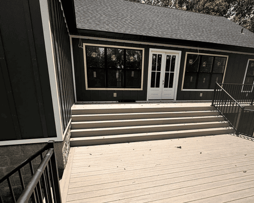 A wide exterior deck with steps leading up to a house with dark siding, white-trimmed windows, and double doors.