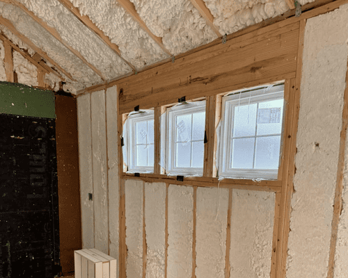 An interior view of a room under construction, showing wooden framing and spray foam insulation around three new windows.