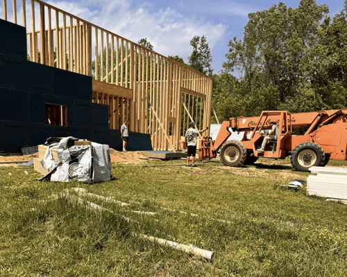 Construction site with a wooden building frame, workers, and an orange telehandler in a grassy field under a blue sky.