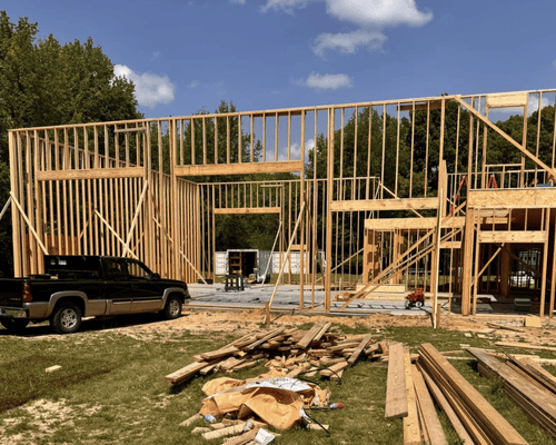 A wood-framed house under construction stands in a grassy lot with a black pickup truck parked in the foreground.