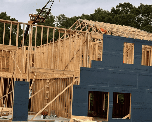 A wood-framed house under construction with grey exterior sheathing, exposed wall studs, and roof trusses against trees.
