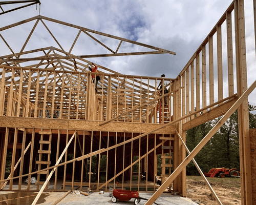 Workers frame the wooden skeleton of a house, with a crane hoisting a roof truss into place on a cloudy day.