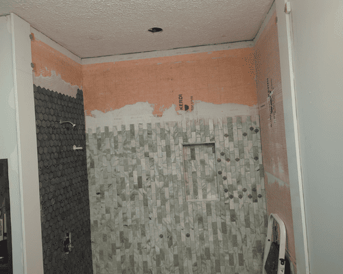 A bathroom under renovation with exposed orange wall backing, dark tile on the left, and gray mosaic tiles on the back wall.