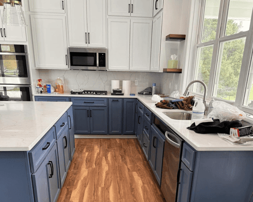 A modern kitchen features white upper cabinets, dark blue lower cabinets, wood flooring, and a white quartz island.