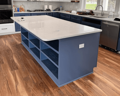 A large, blue kitchen island with open shelving and a white countertop sits on wood-look flooring in a modern kitchen.